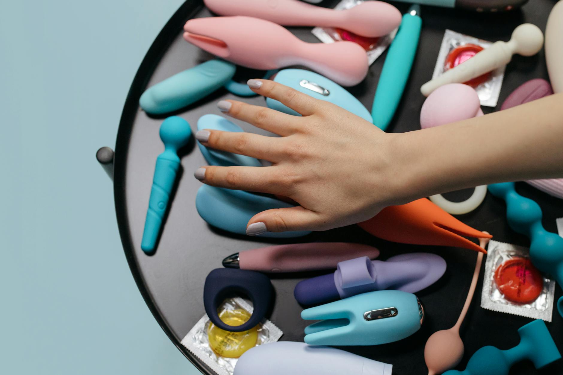 Hand reaching over a collection of colorful vibrators and adult toys arranged together